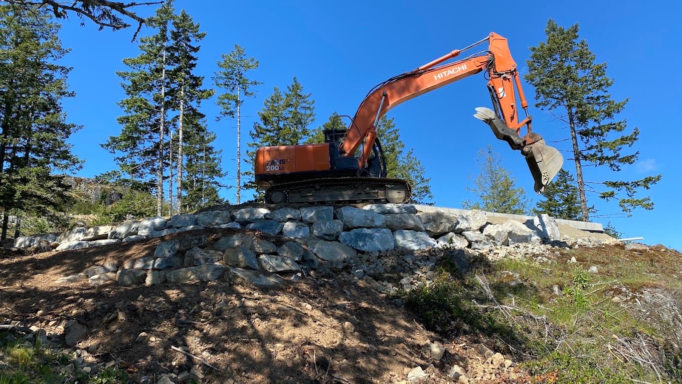 An excavator standing on a finished armour-rock retaining wall against a blue sky