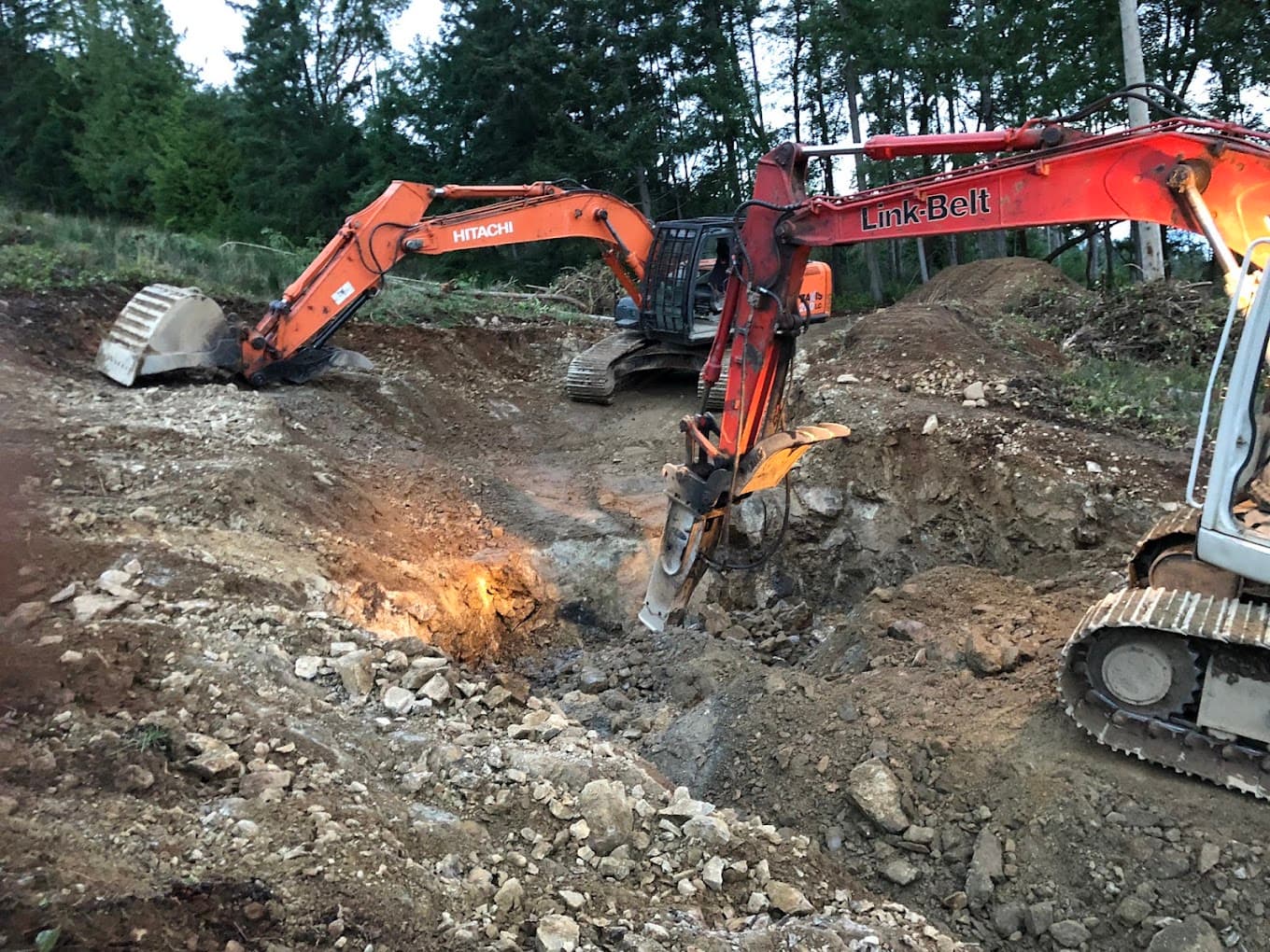 Two excavators, one running a hydraulic hammer on broken rock at dusk