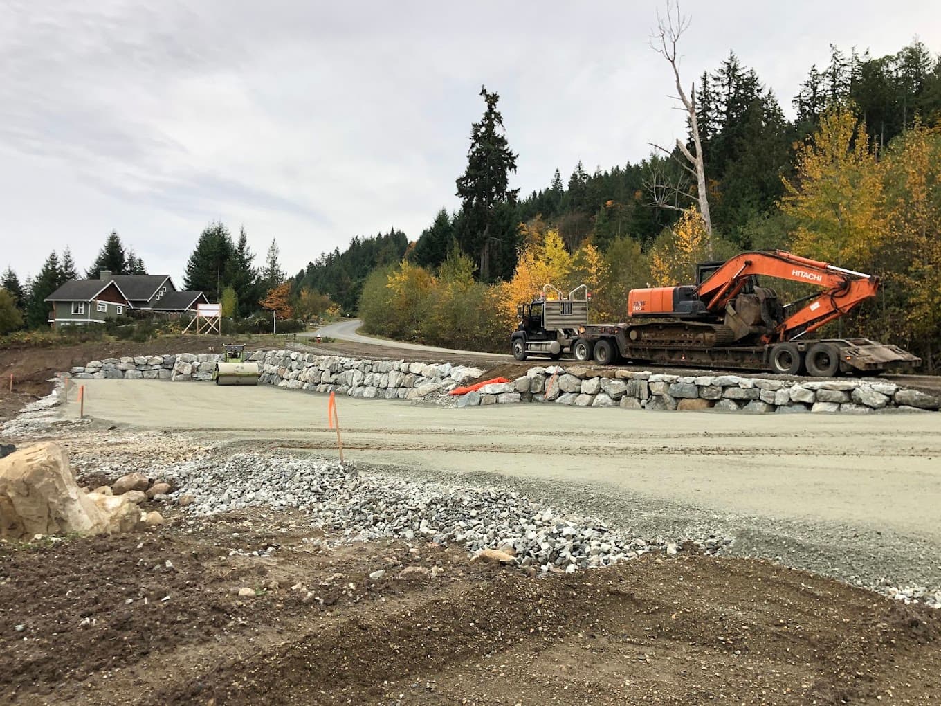 A low-bed truck loaded with an excavator beside a finished rock wall